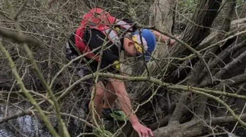 Max Howard The picture shows a person crossing a narrow stream in a wooded area. Branches and tangled undergrowth surround the scene. The person is bent forward, using one hand to steady themselves on a fallen tree trunk that lies across the water like a makeshift bridge. The other hand is reaching ahead for balance.