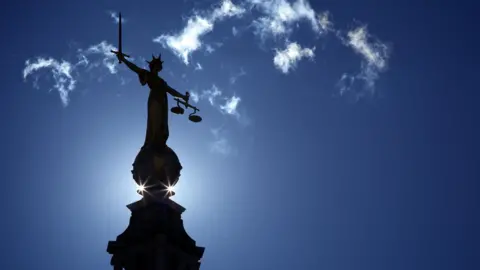 The scales of justice statue above the Old Bailey. The sun rises behind it. There is a blue sky and a few clouds.