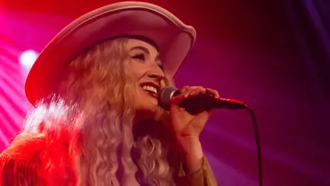 Close up of Janet Devlin singing in front of a hot pink background fading to red. She is holding a mic in her left hand, is smiling and has long wavy blonde hair and a cowboy hat