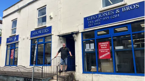A man in shorts and a dark polo shirt stands at the entrance to a white building, with blue shop frontage advertising 'TAPS', 'SHOWERS' and 'GEO JONES & BROS' above three consecutive shop windows. There is a red 'Closing down sale' sign in the main window to the right. 
