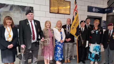 Nine people are standing up near the memorial plaque behind them. One man is carrying a flag with the Royal British Legion named on it.