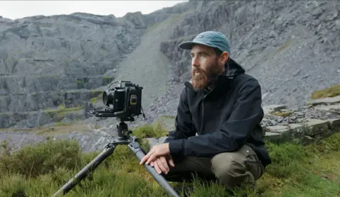 Kyle McDougall Kyle McDougall is wearing a blue baseball hat, navy raincoat, khaki trousers and has a bushy beard. Hi is crouched down on grass in a slate quarry with slate rising up behind him. 