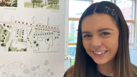 BBC Emily has long hair and is smiling while standing next to some of her design plans on a banner in an exhibition room