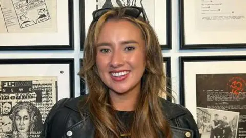 Stacey Burns Stacey Burns, who has long brown hair with blonde highlights stands in front of a series of picture frames on a wall. She has sunglasses on her head and is wearing a black leather jacket.