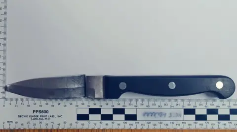 Crown Prosecution Service A black handled kitchen knife, the blade has been snapped away from the hilt but is placed above it. Below is a ruler measuring the blade and handle to be 195cm long