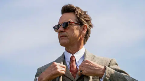 BBC Studios An older man with short hair stands fixing the lapels of the blazer he is wearing. He is dressed very smartly in a white collared shirt and tie, wearing a signet ring on his left hand and a pair of sunglasses. The photo is taken from a slight upward angle looking up to him.