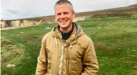 James O'Neill, a man with grey hair, wearing a brown jacket, standing on grass, near a cliff.