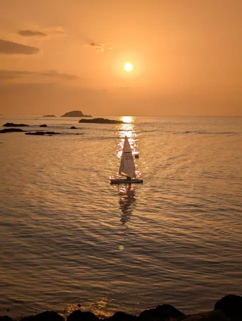 John Welsh An orange sunset over the water. A boat sails below the sun.