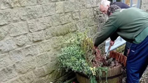 Shaftesbury & District Carers Association Two people, one with white hair and a black jumper using a spade to dig at some plants in a pot - one with a green jumper and blue tracksuit bottoms is pulling at some of the plants