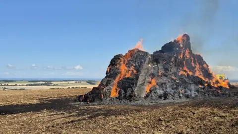 A large mound of blackened hay bales, covered in flames, sits atop a hill. The sky is blue and parched fields can be seen stretching into the distance. 