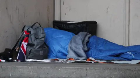 A rough sleeper in a blue sleeping bag sleeps in a doorway with a grey ruck sack by their side.