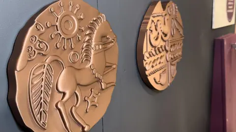 Zoie O'Brien/BBC Two gold coins are placed on a table, one coin has a horse, a leaf and a sun and the second coin has a array of different shapes 