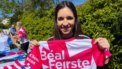 A woman, with long dark brown hair, holding a white and pink jersey reading "Béal Feirste" on the back.