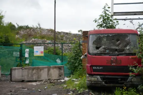 A red truck next to a large waste dump with fencing in front.