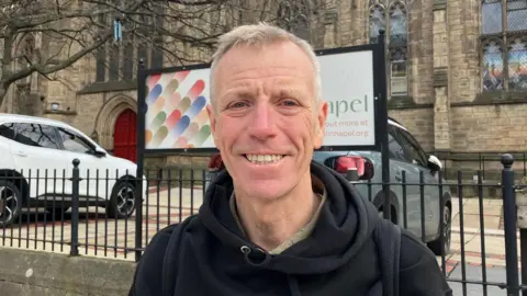 A man with short grey/blonde hair smiles at the camera. He is wearing a black hoodie and standing outside a church.