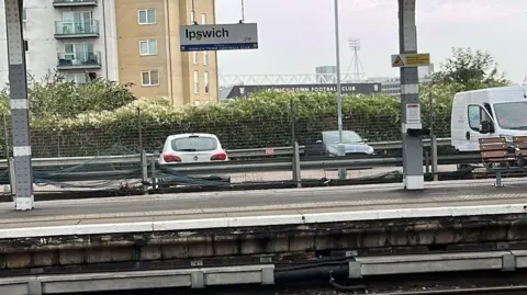 Connor Bennett/BBC An empty platform at Ipswich railway station with an Ipswich sign above the platform, which backs onto a car park with two cars and a van in and Ipswich Town FC ground in the background