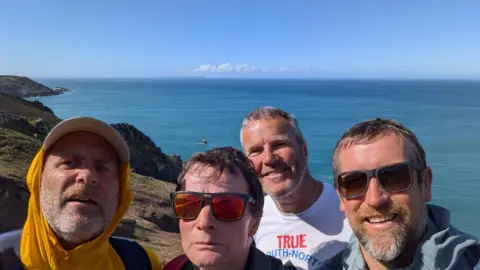 Prof Kevin Dutton (with red sunglasses on) and his three teammates taking a selfie with a coast and sea as background on a sunny day.