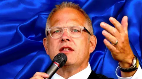 Close up image of Ian Donaghy during a talk. Mr Donaghy is holding a microphone to his mouth. He is holding one hand in the air. 