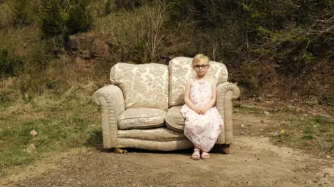 A seven-year-old girl sits on a beige floral sofa, which has been fly-tipped on a piece of land. She has blonde hair, which has been tied back, and a side fringe. She is wearing black glasses, a pink and white floral sleeveless summer dress and a pair of pink sandals. There is gravel around the sofa and some trees and grassland behind it.