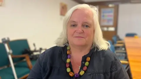 Jemima Laing viste un top negro y un collar de grandes cuentas multicolores y está sentada en una sala de reuniones con un fondo borroso de sillas y una mesa larga.