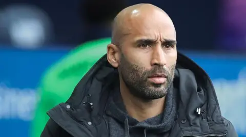 Lee Grant is on the touchline watching on during a match. He is wearing a dark grey coat