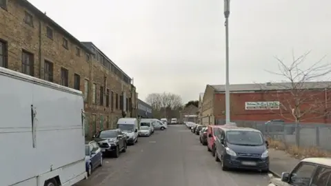 A street in Bradford which has an old mill building on one side and a red brick commercial property on the other. Cars line each side of the street. 