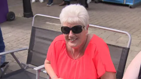 Beth lleva una blusa rosa y gafas de sol. Tiene una cadena plateada alrededor de la mejilla y pendientes de aro. Está sentada en una silla plegable en la calle.