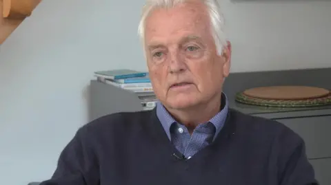 Peter Hall is wearing a dark blue jumper and blue and white checked shirt. He has white hair. There are some books and placemats on the top of a chest of drawers behind him.