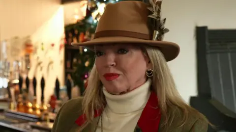 Sue Simmons has brown fedora with a feather in it, white polo neck sweater, long blond hair and a camel coloured jacket. She is in a pub.