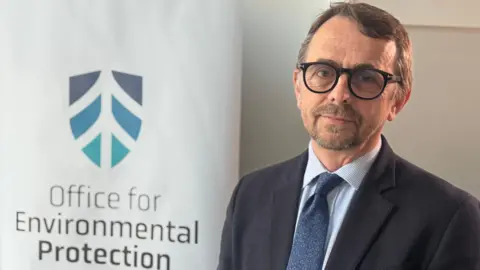 Robbie McDonald is wearing a navy blazer with blue shirt and tie. He has black glasses on. The background is a white poster for the Office For Environmental Protection. 