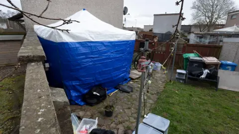 Alan Simpson A blue forensic tent erected in a back garden next to a metal washing line. A number of wheelie bins are in the background and some open plastic boxes 