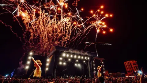 Tom Langford A stage with a large crowd. Screens show there is a performer on the stage, and writing above it reads "Victorious Festival". There are fireworks in the sky.