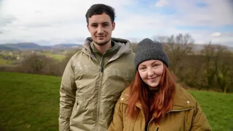 BBC Jade and Jamie are standing in a meadow, side by side, Jade is wearing a beanie hat and Jamie a green coat.