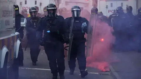 Police officers dressed in all black riot gear, wear black helmets and hold clear riot shields. Behind them are three white police vans, a red flare gas gone off beside them, it emits red smoke. 
