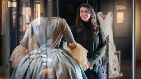 Historic Royal Palaces Claudia Acott Williams looking at one of the dresses in a glass cabinet. She has long brown hair, wearing a dark green jumper with her hands folded together. The dress is a Georgian-style costume gown in blue with white lace on the sleeves. The coronation toile can be seen in background. It is a white gown with gold embroidery.