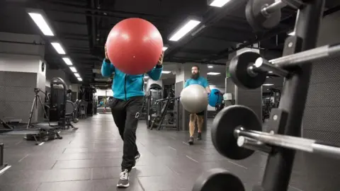 A woman face is obscured by an exercise ball she is carrying past workout equipment in an indoor gym.