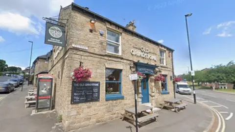 Google Exterior of The Old Grindstone pub, a stone-built corner building with blue-painted doors and window frames, hanging flower baskets, outdoor picnic benches and prominent pub signage.