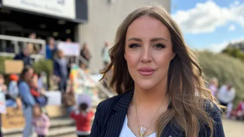 Lindsay Love - a woman with long brunette hair and a dark jacket on - looks to the camera while standing outside a nursery. Out of focus children and parents are in the background.