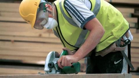 A young man wearing a hard hat and goggles sawing a piece of wood using a circular saw