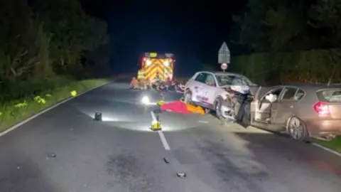 A road with an ambulance and two crashed cars. A white car and black car have hit each other head-on.