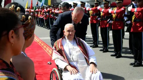 Getty Images Paus Franciscus wordt in een rolstoel over de rode loper op de luchthaven van Dili geduwd. Links van hem staat een rij soldaten in rode uniformen.