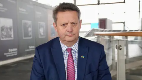Alun Davies MS is wearing a suit and tie will posing for a photograph in the Senedd. He is wearing a royal blue suit with a white and blue checked shirt and a pink tie with a blue pattern on 
