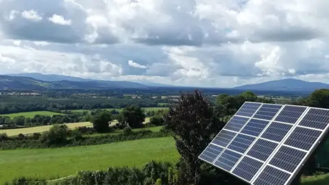 Michael Wilkinson A board of solar panels set on a sloped garden outside the Wilkinson family's home on a sunny day in County Kilkenny. The board can be swivelled to point the panels at the sun's rays.  The garden is surrounded by the green fields of the Suir Valley with hills in the distance.