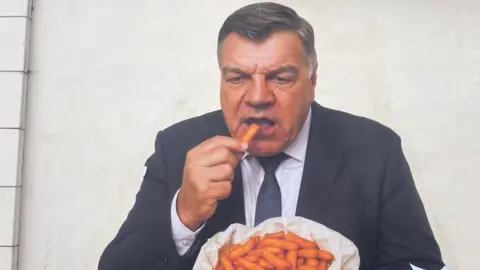 Sam Allardyce is painted on the side of a white wall wearing a dark navy blue suit eating orange chips out of a white paper bag. 