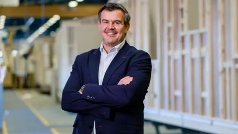 Rix Group/Neil Holmes A man standing inside a factory wearing a blue suit jacket and smiling. There are several static caravan frames in the background.