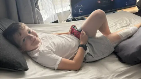 Nine-year-old Theo Rees lying in bed at home holding a red Xbox remote. He is wearing a white t-shirt and grey jogging shorts and has a big plaster on his right leg.