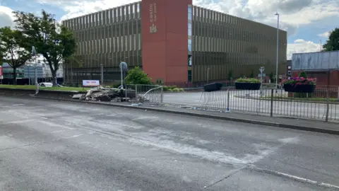 A street scene shows a damaged brick wall and police tape, suggesting a recent crash. A tree and the "Castle Car Park" can be seen in the background.
