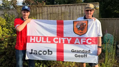 Jasmine Lowe/BBC News Jacob and Geoff are stood in a garden in front of a bush. They're holding a large England flag, but on the top left of the flag there's a Hull City emblem. On the red cross it says 'Hull City AFC' and either side of the bottom corner it reads Jacob and Grandad Geoff. 