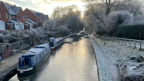 John Sadler Boats are moored on the canal at Gnosall. The verges next to the canal are covered in frost and there is ice on the water. A row of houses stands next to the canal.