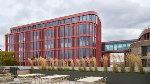External view of the Forum in Gloucester. The building has red steel cladding between windows. The foreground shows an outdoor terrace surrounded by a grey planter with grasses in. 